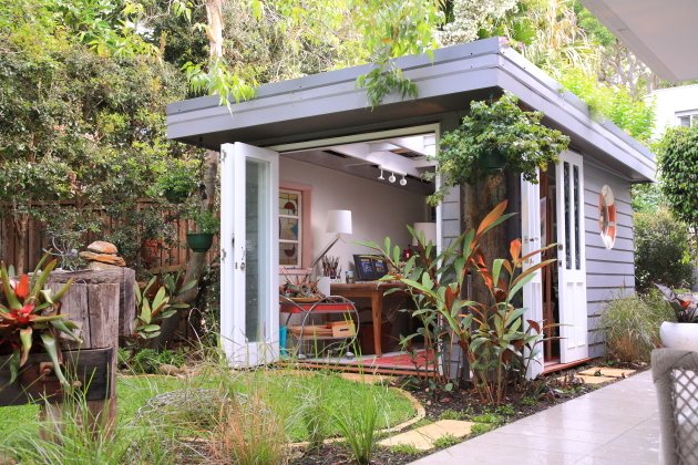 Grey Shed in a lush garden field backyard. It has double doors and looks like a creative space for someone to work in. 